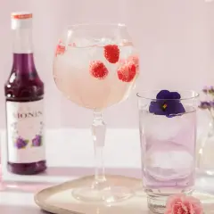 Dwa drinki z malinami i fiołkiem, obok buteleczki syropów Monin.