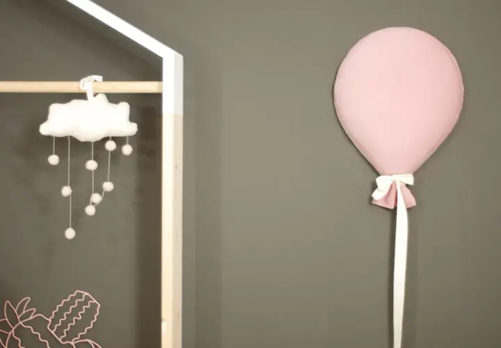 Różowy balon z kokardką przyczepiony do szarej ściany obok dziecięcej dekoracji.
