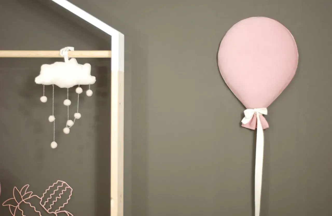 Różowy balon z kokardką przyczepiony do szarej ściany obok dziecięcej dekoracji.
